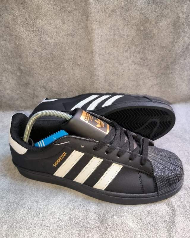 Adidas Adidas Superstar (sepatu lifestyle/casual klasik dengan ciri khas pelindung jemari berbahan karet bertekstur tempurung (shell-toe), logo tiga garis khas Adidas di samping, serta cetakan tulisan emas 